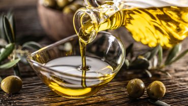 Pouring olive oil from carafe to the glass bowl and in background are olive branches put on table.