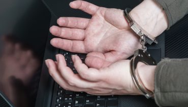 Arrested hacker with hands in handcuffs on the laptop keyboard and digital code around. Cyber crime concept.